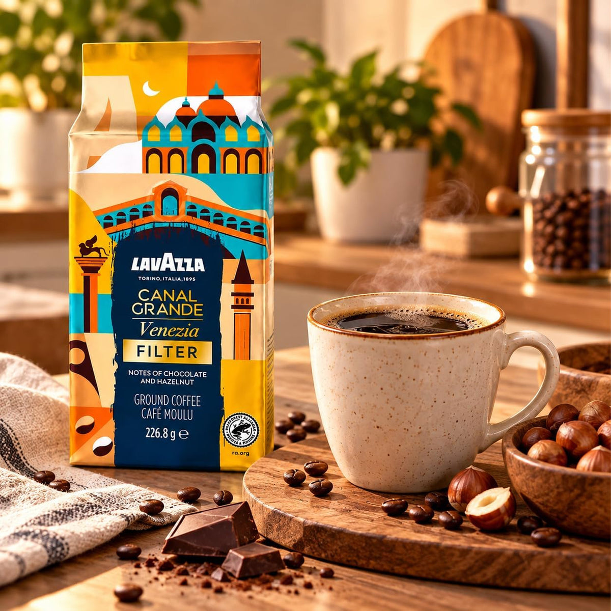 Lavazza Venezia Canal Grande Filter coffee in a colorful illustrated bag, displayed next to a steaming cup of coffee on a wooden table with chocolate, nuts, and plants in a warm kitchen setting.
