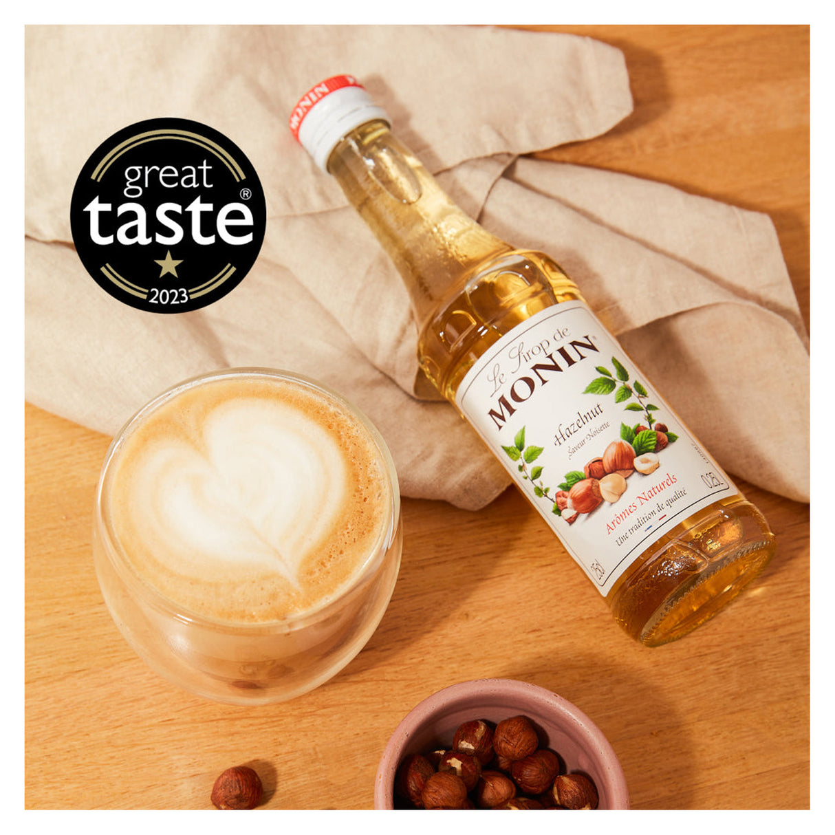Monin hazelnut syrup bottle with a cup of coffee and nuts on a wooden surface, featuring the Great Taste 2023 award.
