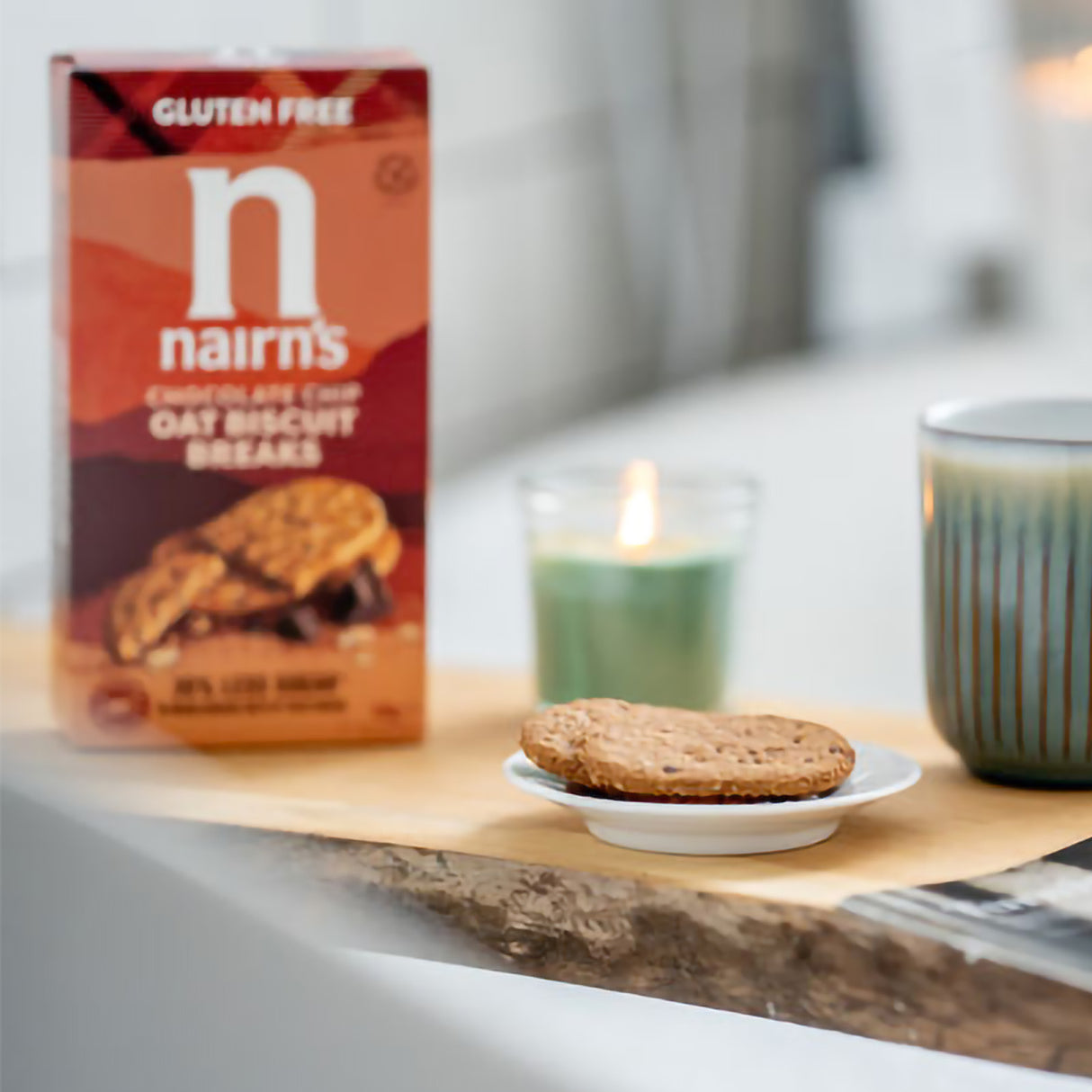 Nairn's gluten-free oat biscuit breaks box with a cookie on a plate, candle, and mug in the background.