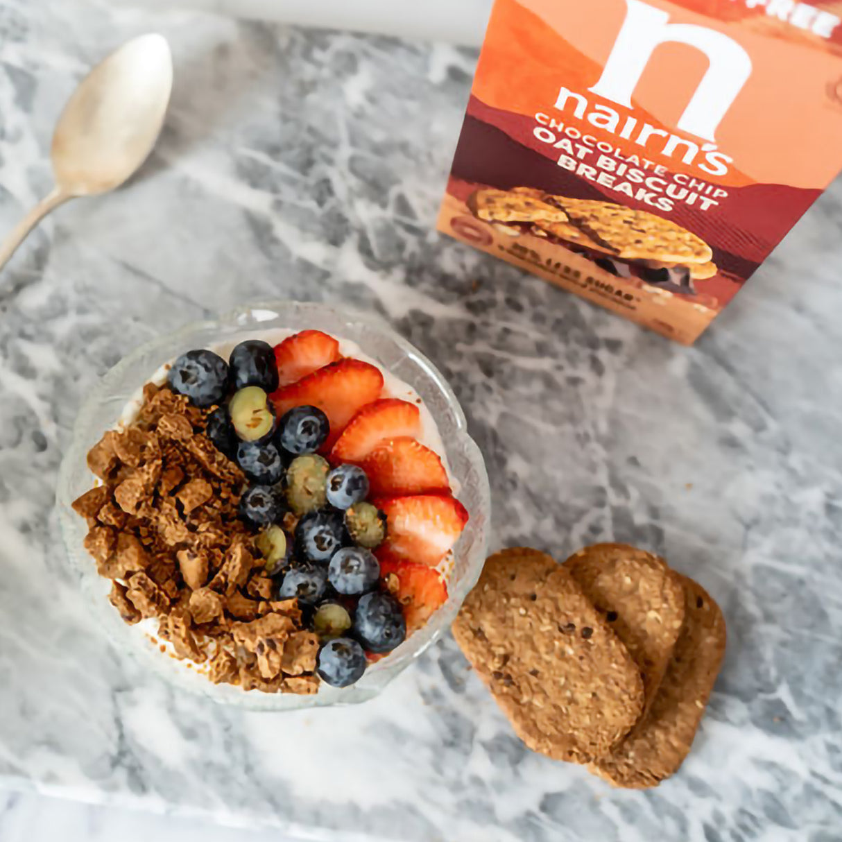 Yogurt bowl with granola, berries, and a box of Nairn's chocolate chip oat biscuits on a marble surface.
