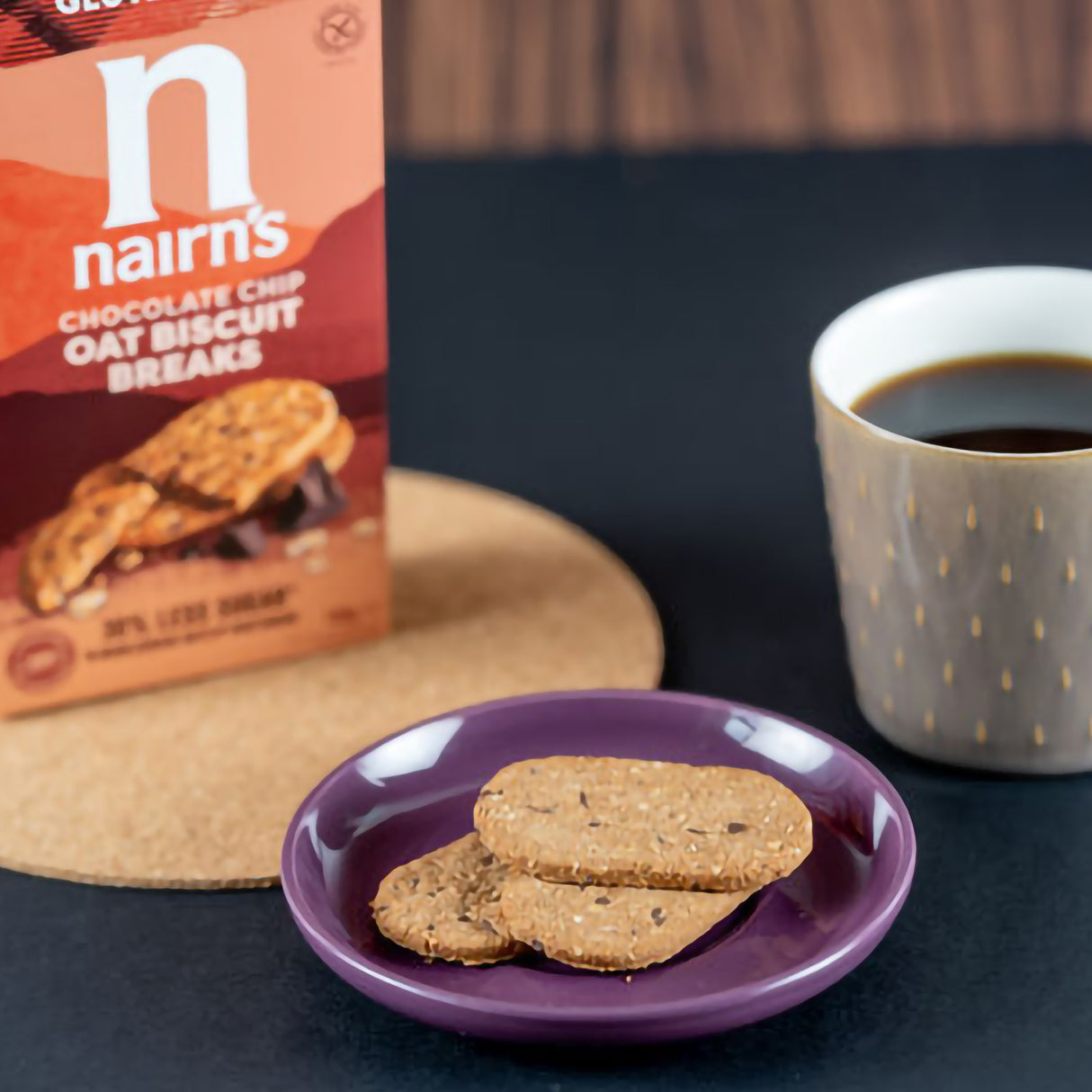 Nairn's chocolate chip oat biscuit breaks with a box and a cup of coffee on a dark surface.