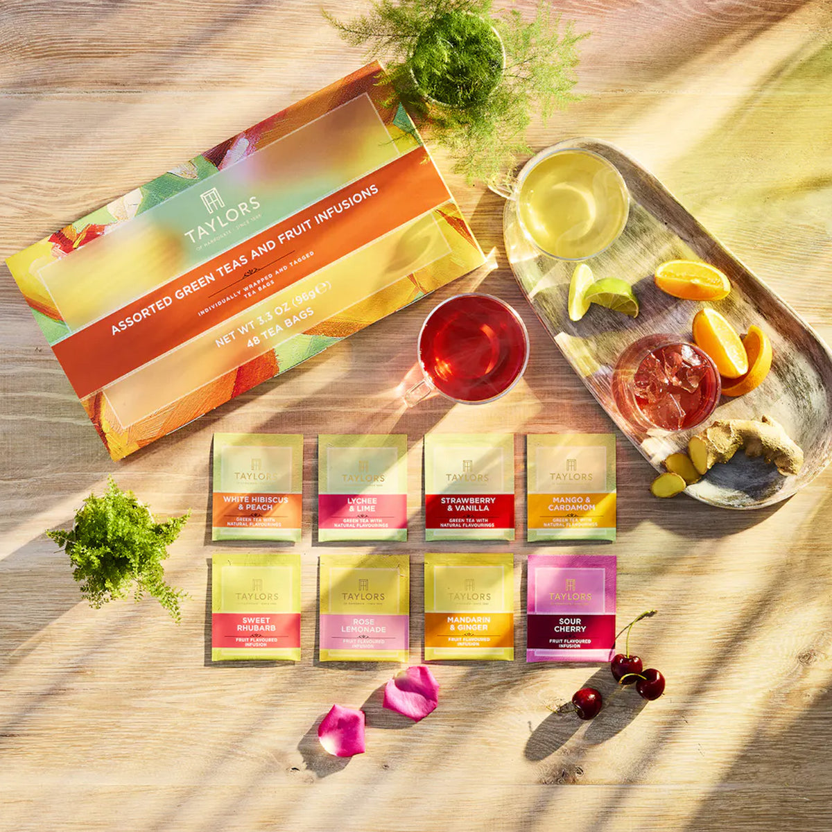 A styled flat-lay of Tailors assorted green tea and fruit infusions tea bags, showing the colorful retail box, individual sachets, brewed tea in cups, and fruit garnishes on a sunlit wooden surface.