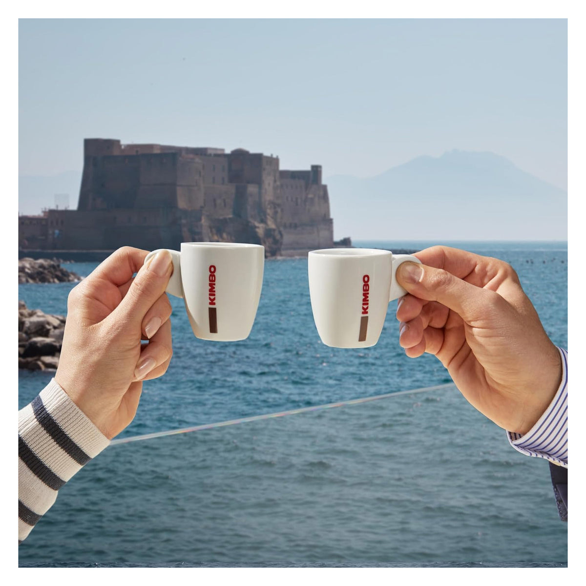 Two hands holding white ceramic espresso cups with red KIMBO branding, posed in front of a scenic background of a coastal fortress and blue sea.