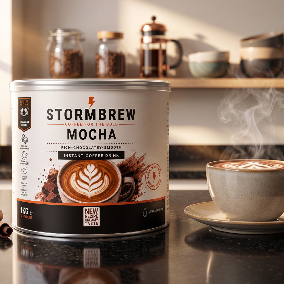 1kg tin of Stormbrew Mocha instant coffee drink sits on a dark countertop beside a steaming cup of mocha latte, with coffee jars and a French press blurred in the background.