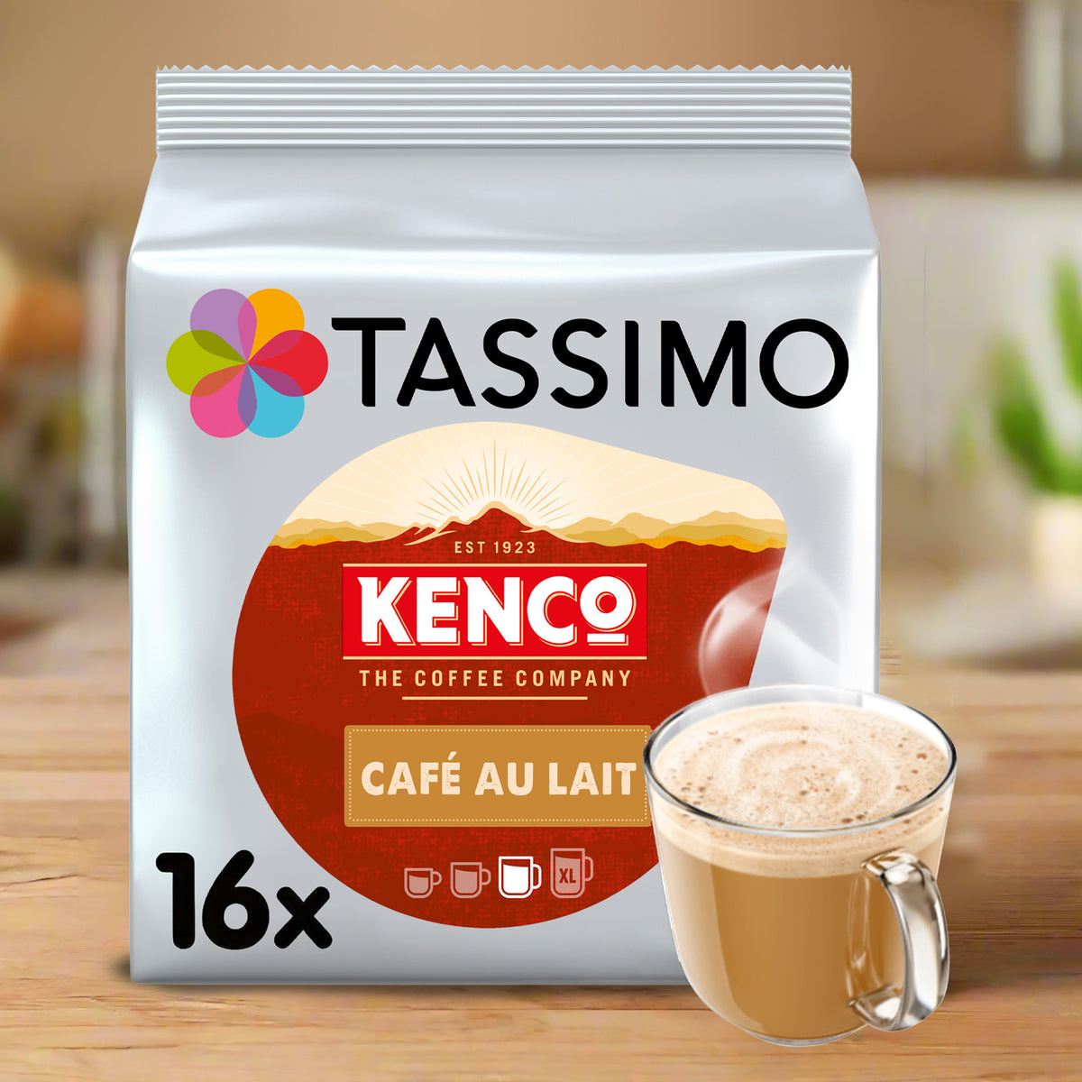 A package of Tassimo Kenco Café Au Lait coffee pods shown on a wooden surface with a glass mug of latte in front. The bag is white with a large red circular logo and multicolor flower icon, and the mug contains a light brown coffee drink.