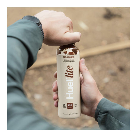 Hand holding a Huel Lite chocolate protein shake bottle against a blurred natural background