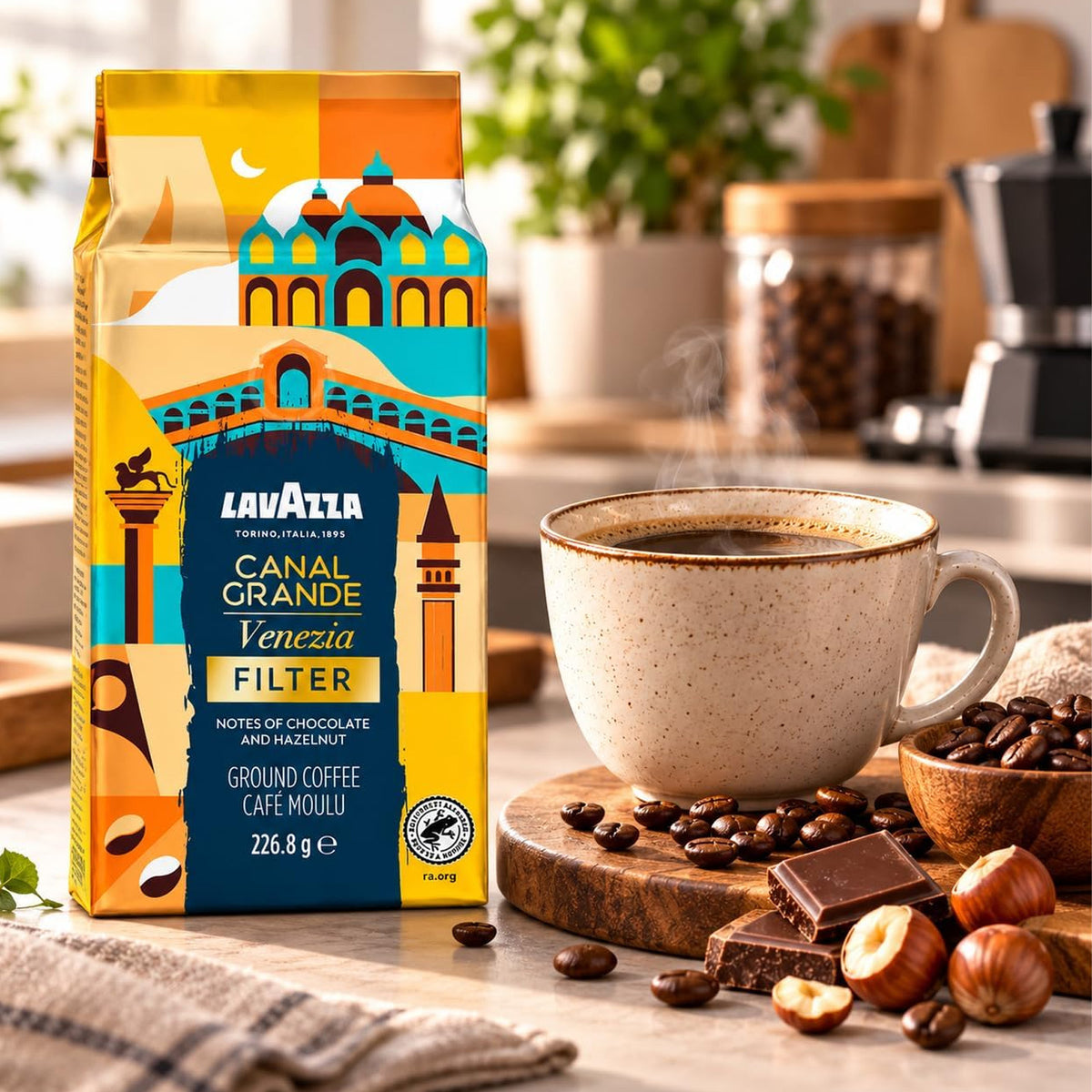 Lavazza Venezia Filter coffee bag standing on a kitchen counter beside a ceramic cup of coffee, coffee beans, chocolate, and hazelnuts, with a softly blurred kitchen background.