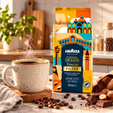 Lavazza Canal Grande Venezia filter coffee in a colorful illustrated bag, styled on a kitchen counter beside a steaming cup of coffee, scattered coffee beans, chocolate pieces, and a wooden spoon of ground coffee.