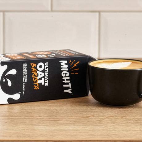 Mighty Oat Barista packaging with a black mug of oat milk latte on a wooden surface.