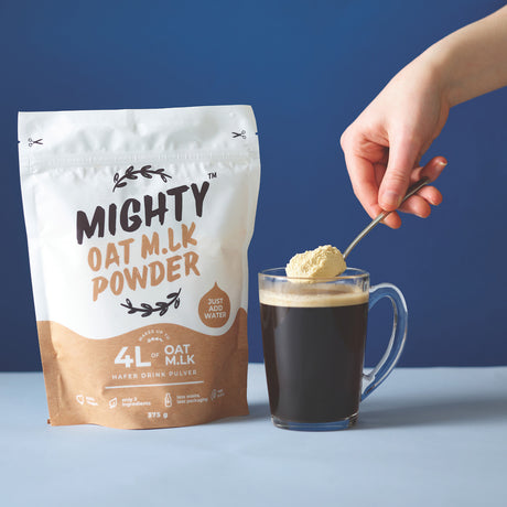 A hand holds a spoon adding oat milk powder to a glass of oat milk, next to a bag of Mighty Oat Milk Powder on a light blue surface against a dark blue background.