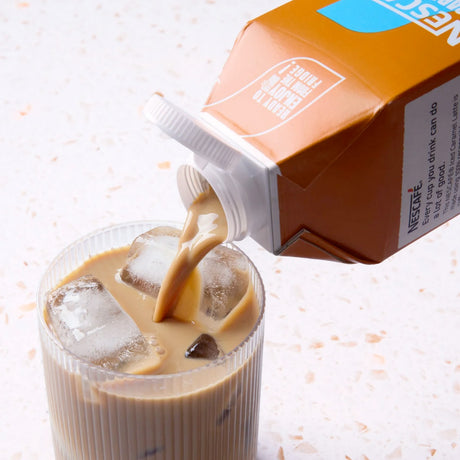 Nescafe instant coffee being poured into a glass with ice cubes on a light background