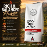 A bag of Northern Beaches Wood Fired Coffee Beans stands on a dark forest-themed background with flavor text and tasting notes. The bag is white with black lettering, a red band reading COFFEE BEANS, and a small flame logo near the bottom.