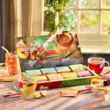 A boxed tea gift set displayed on a wooden table in a bright kitchen or tea room setting, with cups of tea, glasses, and tea bags arranged around the open package. The box prominently reads 'Tastefully British' and shows multiple colorful tea packet designs.