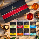 Taylors Assorted Speciality Teas gift set displayed on a wooden surface with two cups of tea, decorative ribbons, a candle, dried citrus slices, and small star-shaped ornaments. The set includes a large black and red box and eight individual tea bags with colorful labels.
