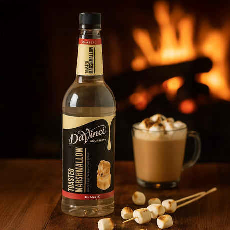 Bottle of DaVinci Gourmet toasted marshmallow syrup with a cup of hot chocolate and marshmallows in front of a fireplace.