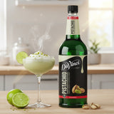 Bottle of DaVinci Gourmet Pistachio syrup with a cocktail and limes on a wooden surface.