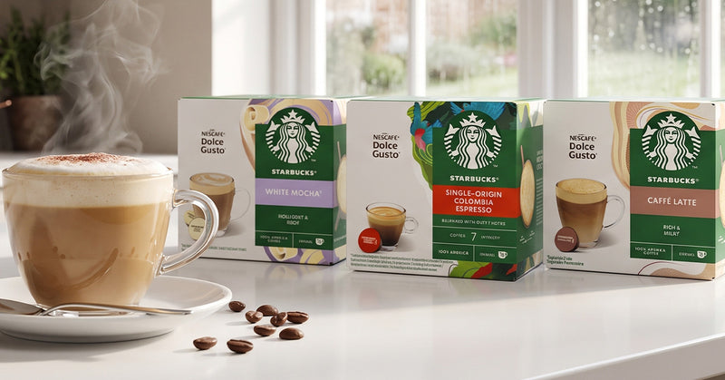 Four Starbucks coffee capsule boxes lined up on a white countertop next to a steaming cup of cappuccino, with coffee beans scattered in front and a softly blurred kitchen background.