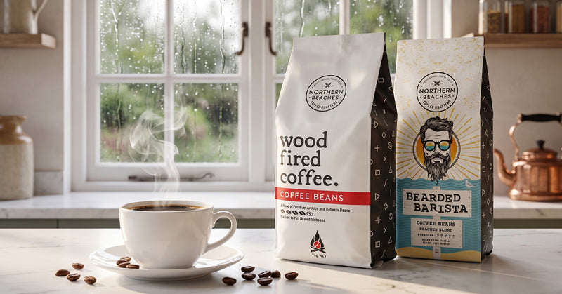 Two bags of Northern Reach Coffee beans on a kitchen counter next to a steaming cup of coffee in front of a rainy window.