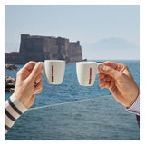 Two hands holding white ceramic espresso cups with red KIMBO branding, posed in front of a scenic background of a coastal fortress and blue sea.