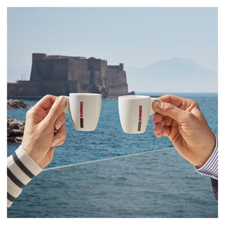 Two hands holding white ceramic espresso cups with red KIMBO branding, posed in front of a scenic background of a coastal fortress and blue sea.