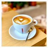 A white ceramic cup of coffee with latte art sits on a matching saucer with a spoon, placed on a light wooden table in a softly blurred indoor setting. The cup has a visible red KUMBO logo.