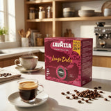 A box of Lavazza Lungo Dolce coffee capsules sits on a kitchen counter next to two cups of coffee, with scattered coffee beans in the foreground and a warm kitchen interior in the background.