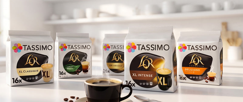 Five Tassimo coffee pods packages in different flavors arranged on a white countertop with a cup of coffee and coffee beans in front, against a softly blurred kitchen background.