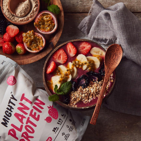 Fruit bowl with a wooden spoon, surrounded by ingredients and a 'Mighty Plant Protein' bag on a wooden surface.