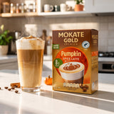 A box of Mokate Gold Premium Pumpkin Spice Latte coffee stands on a kitchen counter beside a tall glass of iced pumpkin latte with whipped cream and cinnamon. The scene is set in a warm, softly blurred kitchen with coffee mugs on a shelf in the background.