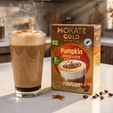 A box of Mokate Gold Premium Pumpkin Spice Latte coffee sachets stands next to a tall glass of prepared pumpkin spice latte with foam and cocoa dusting, on a light countertop with coffee beans scattered nearby.
