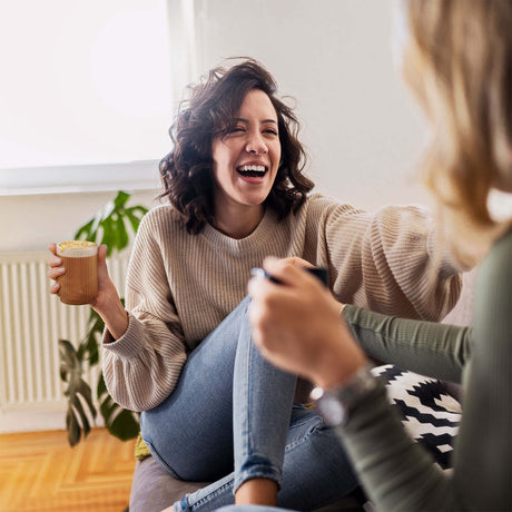 Two women sitting on a couch, one holding a drink and laughing.