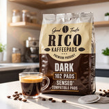 A bag of Rico Kaffepads Dark coffee filters stands on a kitchen counter next to a steaming glass of espresso, with coffee beans scattered around and Senseo-compatible pads in front. The background shows a softly blurred kitchen setting.