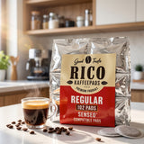 A bag of Rico Kaffepads Regular coffee filters stands on a kitchen counter beside a glass of espresso, with coffee beans scattered around and a blurred kitchen background.