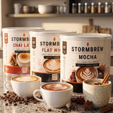 Three Stormbrew instant coffee cans labelled Chai Latte, Flat White, and Mocha, displayed on a counter-top with matching coffee cups, coffee beans, and spices in a kitchen setting.