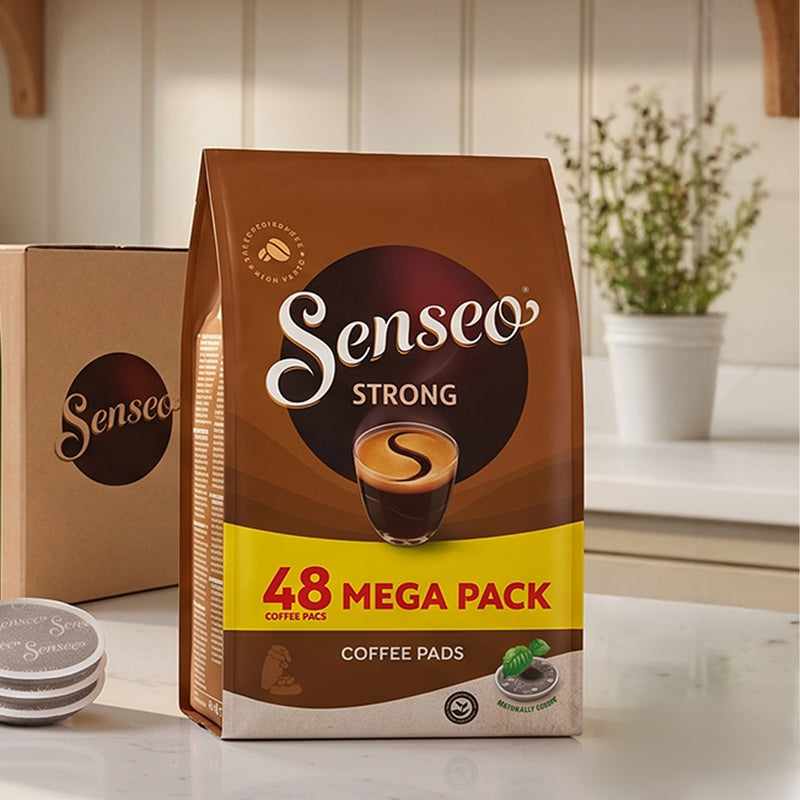A Senseo Strong 48 Mega Pack coffee pad bag stands on a countertop in a bright kitchen setting, with a matching Senseo box and a small stack of coffee pads visible beside it.