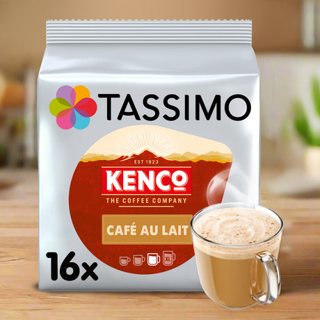 A package of Tassimo Kenco Café Au Lait coffee pods shown on a wooden surface with a glass mug of latte in front. The bag is white with a large red circular logo and multicolor flower icon, and the mug contains a light brown coffee drink.