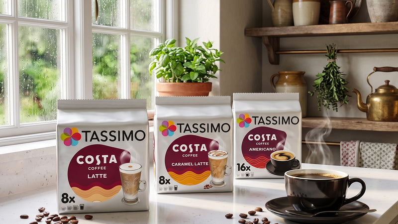 Three packs of Tassimo Costa coffee pods arranged on a kitchen counter by a window, with a cup of coffee and scattered coffee beans in a real kitchen setting.