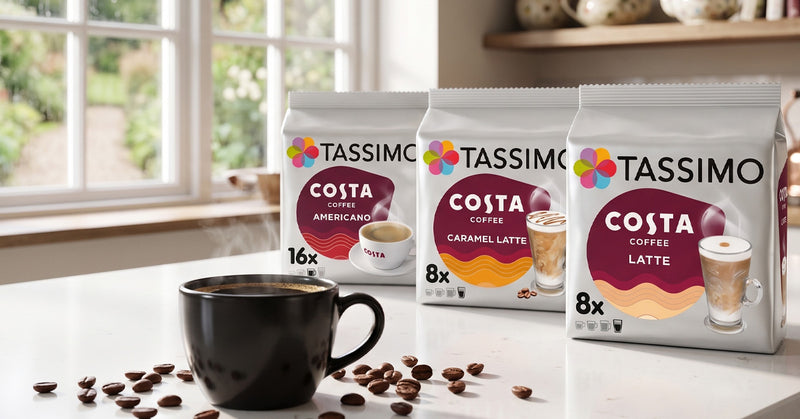 Three bags of Tassimo Costa Coffee beans (Americano, Caramel Latte, and Latte) are displayed on a white countertop next to a black coffee mug with scattered coffee beans, set against a bright kitchen background with a window.