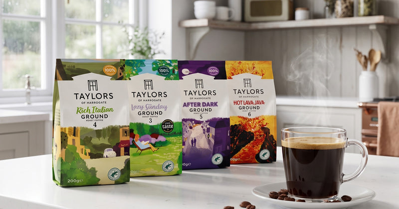 Four bags of Taylors of Penzance coffee beans in different flavors and roast levels, displayed on a white countertop next to a glass mug of freshly brewed black coffee with a few coffee beans scattered on the saucer. The background shows a softly blurred kitchen interior.