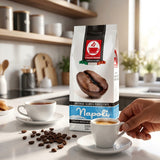 A bag of Napoli espresso coffee stands on a kitchen counter with two white cups, one filled with espresso and one being held by a hand. Coffee beans are scattered on the counter, with a softly blurred kitchen background.