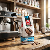 A bag of Napoli coffee beans stands on a rustic wooden counter in a cafe setting, with an espresso machine on the left and shelves of coffee supplies in the background.