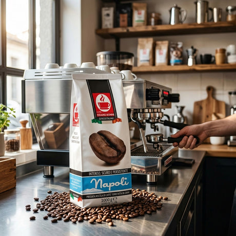 A bag of Napoli espresso beans stands on a stainless steel counter in a coffee shop, with coffee beans scattered around the base and a person's hand operating an espresso machine in the background.