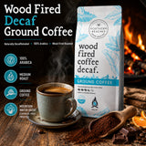Northern Beaches Wood Fired Decaf Ground Coffee bag shown on a rustic wooden table with a cup of coffee, coffee beans, chocolate, and orange slices, alongside text callouts and a warm fire background.