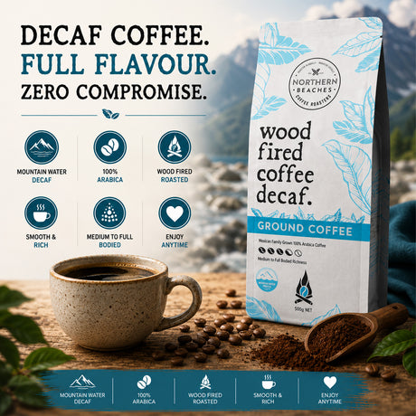 Northern Beaches Wood Fired Coffee Decaf Ground Coffee stands on a rustic wooden surface next to a cup of black coffee, scattered coffee beans, and a wooden spoon of ground coffee. The background features a scenic mountain view with a blue sky, and the top and bottom of the image feature feature informational text and icons detailing the coffee's attributes.