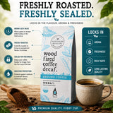 Wood Fired Coffee Decaf ground coffee is shown on a rustic wooden surface with a ceramic coffee cup, coffee beans, and a wooden scoop. The image includes marketing text highlighting freshness, aroma lock valve technology, and benefits of wood fired roasting.