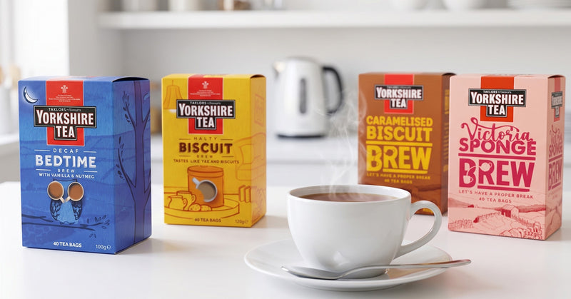 Four Yorkshire Tea boxes arranged on a white countertop beside a white teacup and saucer with a spoon, with a blurred kettle and kitchen background.